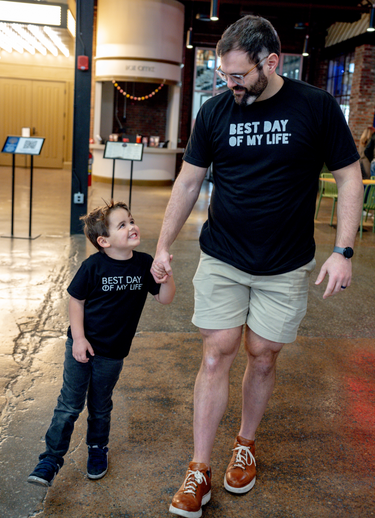 white child wearing black t-shirt with BEST DAY OF MY LIFE written in white smiling up at white man with glasses also wearing black crewneck t-shirt with best day of my life written across in white